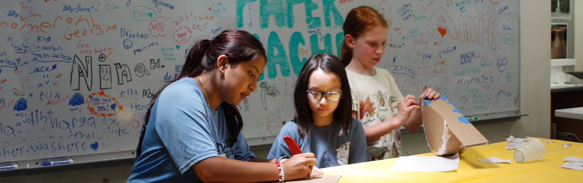 Staff shows a girl how to cut the carboard, while another student puts together hers